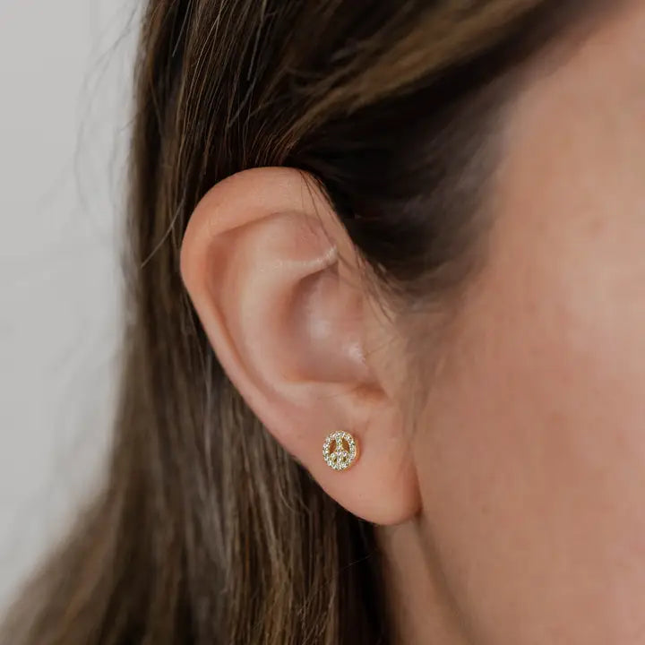 Close-up of a woman's ear with brown hair, wearing JaxKelly's Pavé Peace Sign earring—a small, circular gold stud adorned with sparkling CZ stones and finished in 18k gold plating. The softly blurred background highlights its elegance.
