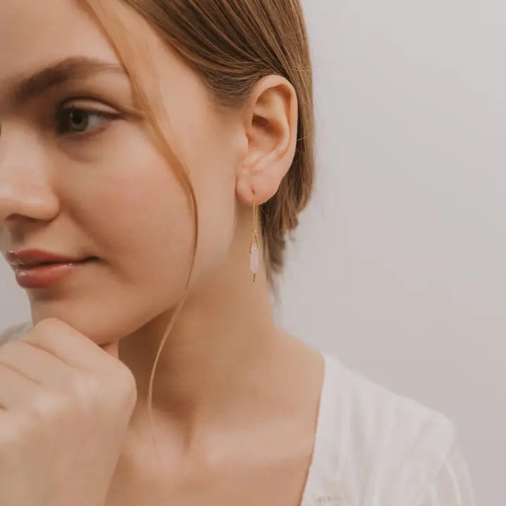 A young woman with light skin and brown hair wears a white top and JaxKelly's Gemstone Threader - Rose Quartz earrings. She gazes left, hand near her chin, highlighting the minimalist style of this elegant jewelry against a plain background.