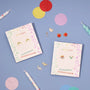 Two JaxKelly Dinosaur Enamel Flat Back Kids Earring sets, featuring playful, colorful designs like dinosaurs, rainbows, apples, and toast—perfect for sensitive ears. Blue surface with scattered candles and confetti adds a festive touch.