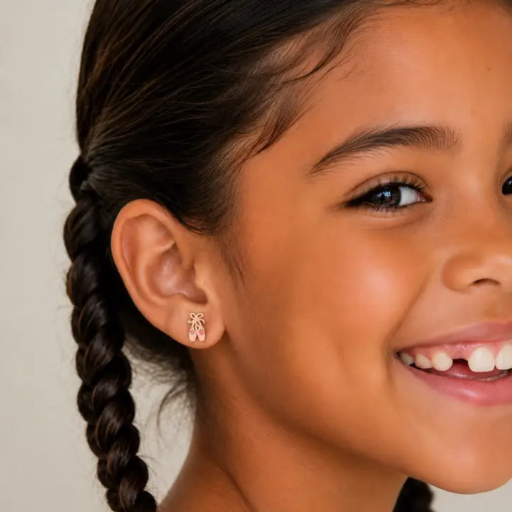 Close-up of a smiling young girl with dark braided hair, wearing JaxKelly Ballet Slippers Enamel Flat Back Kids Earrings—small, gold, hypoallergenic studs designed for sensitive ears—against a neutral background.