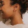A person with curly hair smiles gently, wearing JaxKelly's Gemstone Threader - Smoky Quartz earring with a geometric pendant, set against a light background.
