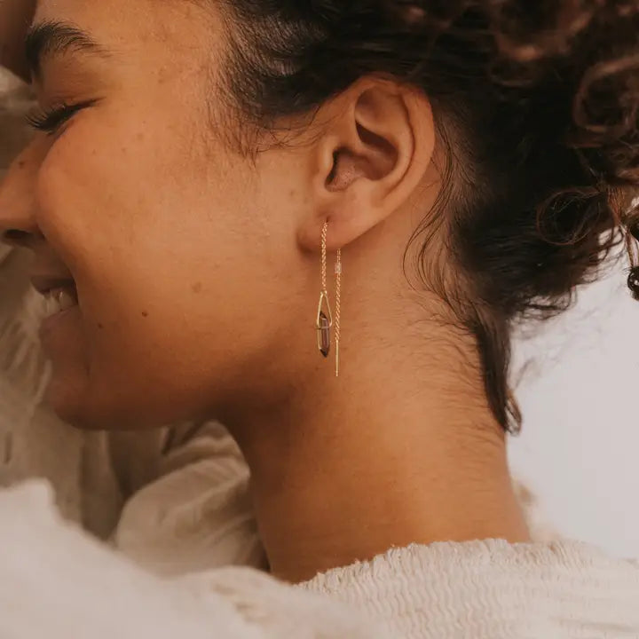 A person with curly hair smiles gently, wearing JaxKelly's Gemstone Threader - Smoky Quartz earring with a geometric pendant, set against a light background.