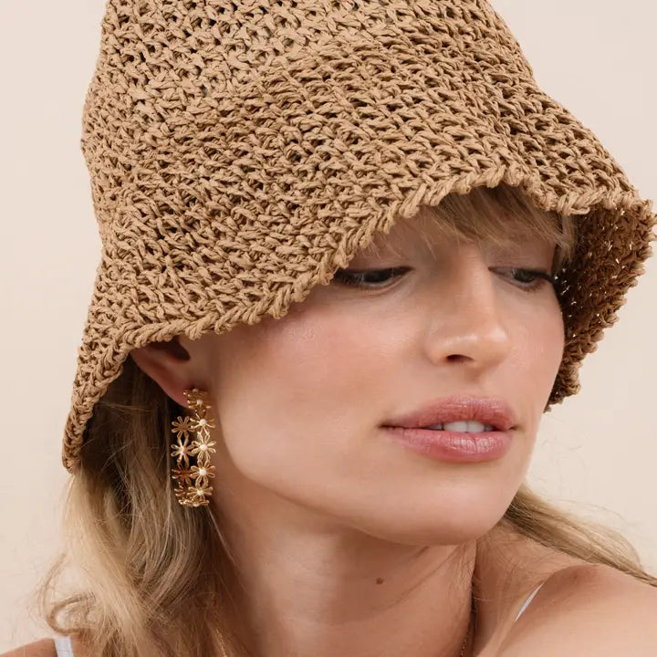 A woman with light blonde hair and natural makeup wears a tan woven bucket hat and JaxKelly Daisy Chain Earrings – Enamel and Gold, gazing slightly downward.