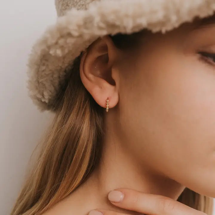 A person with long light brown hair wears a beige fuzzy hat and JaxKelly Small Ball Hoop earrings. Their hand rests gently on their neck against a plain, light-colored wall.