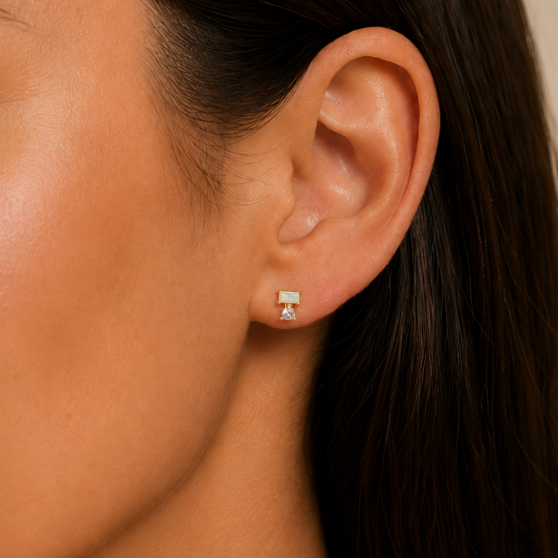 Close-up of a woman’s ear with straight brown hair, wearing the JaxKelly Double Stud Stack - White Opal: a small, rectangular white opal stud earring crafted from hypoallergenic, surgical grade stainless steel, featuring a delicate dangling charm underneath. The neutral background keeps the focus on the earring.