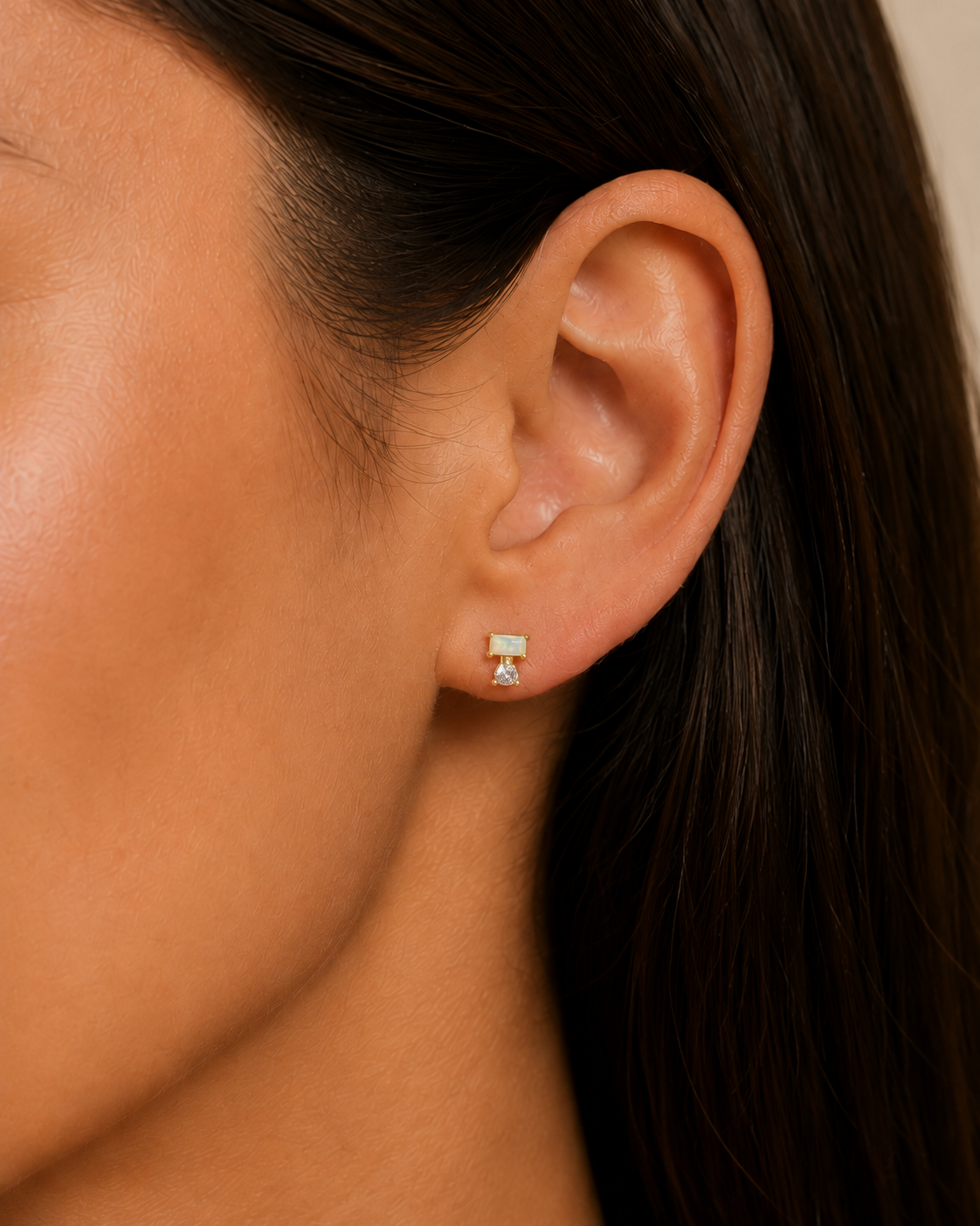 Close-up of a woman’s ear with straight brown hair, wearing the JaxKelly Double Stud Stack - White Opal: a small, rectangular white opal stud earring crafted from hypoallergenic, surgical grade stainless steel, featuring a delicate dangling charm underneath. The neutral background keeps the focus on the earring.