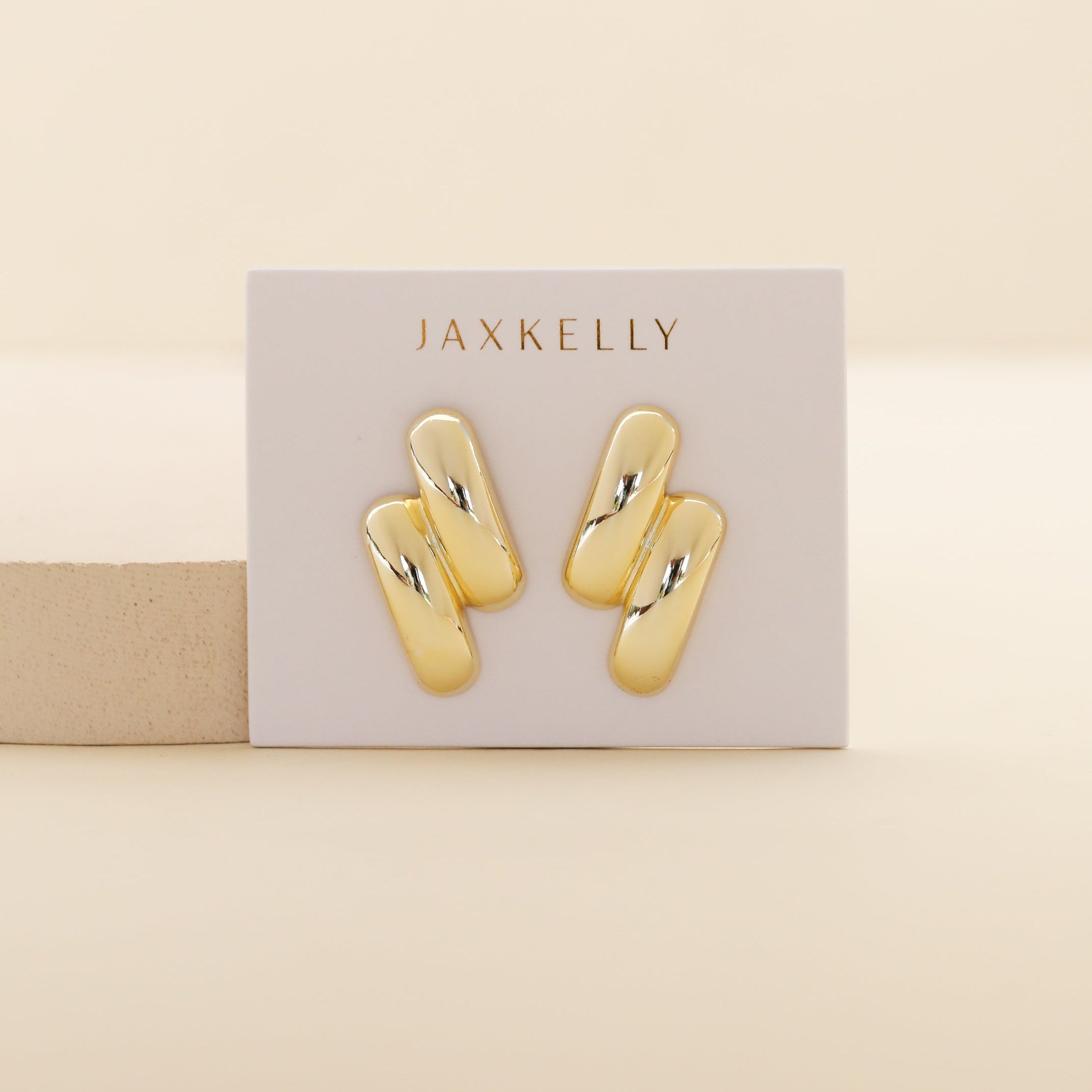 A white card labeled JaxKelly displays two pairs of modern Gold Double Bold Bar stud earrings with shiny, curved bars and 18k gold plating against a light beige background.