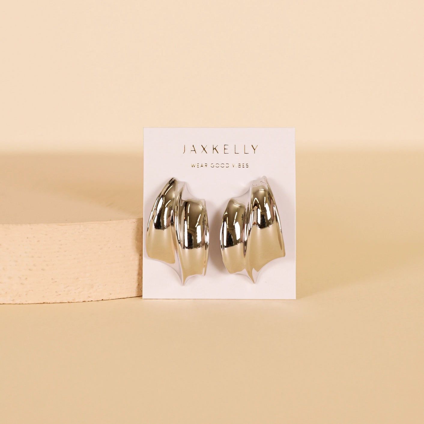 The Bold Wave - Silver Earrings by JaxKelly feature a shiny, wave-inspired design and sterling silver posts, displayed on a white JaxKelly card with a beige background and a textured block on the left side.