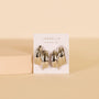 The JaxKelly Silver Bold Wave statement stud earrings, featuring shiny silver waves and sterling silver posts, are displayed on a white branded card against a neutral beige background with a textured block beside them.