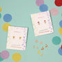 A pair of Cherry Enamel Flat Back Kids Earrings for sensitive ears on decorative cards, displayed on a teal surface with colorful confetti. These hypoallergenic earrings feature playful cherry designs.
