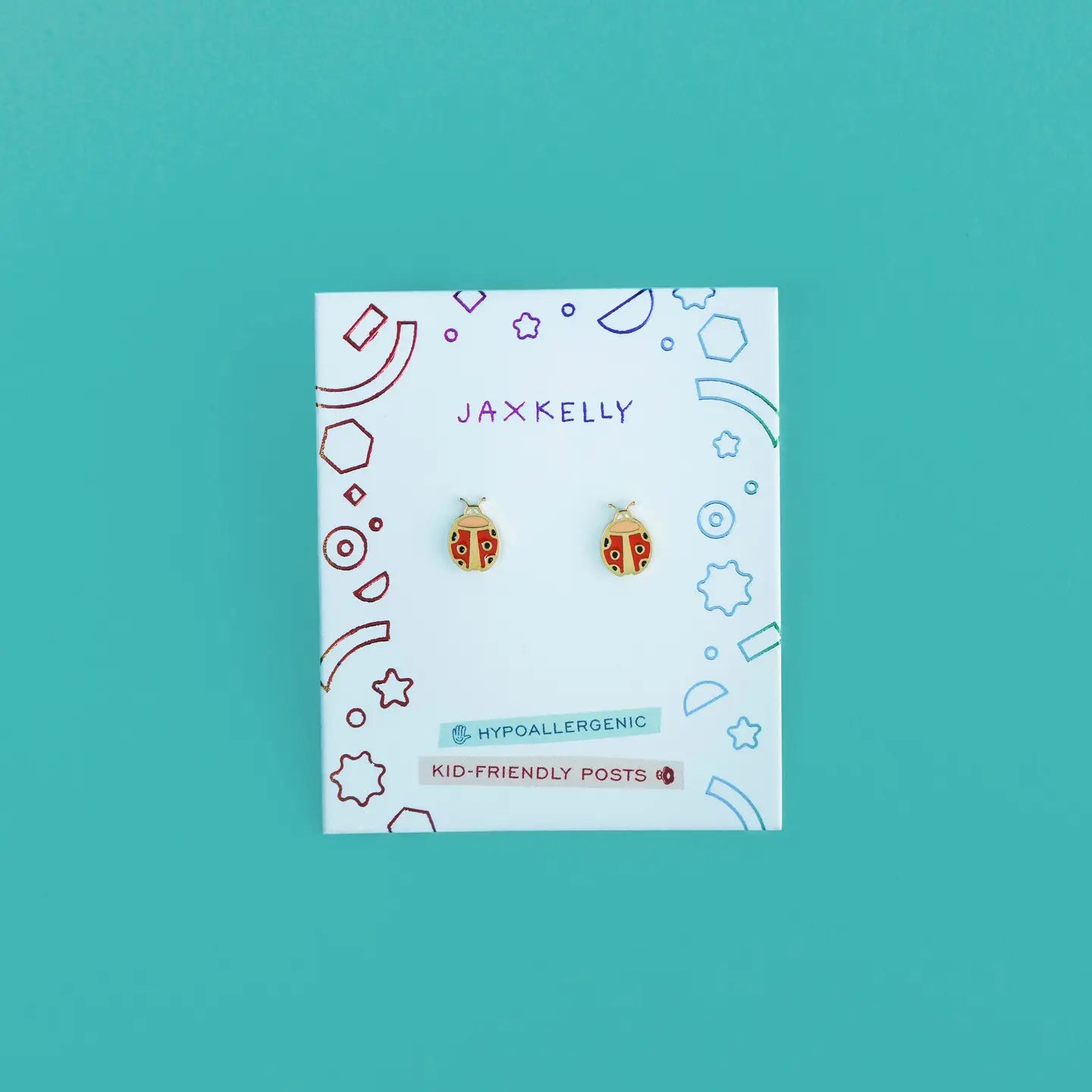 A pair of Ladybug Enamel Flat Back Kids Earrings in red and gold, hypoallergenic and safe for sensitive ears, shown on a white JaxKelly card with colorful geometric designs against a teal background.