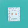 A pair of Ladybug Enamel Flat Back Kids Earrings in red and gold, hypoallergenic and safe for sensitive ears, shown on a white JaxKelly card with colorful geometric designs against a teal background.