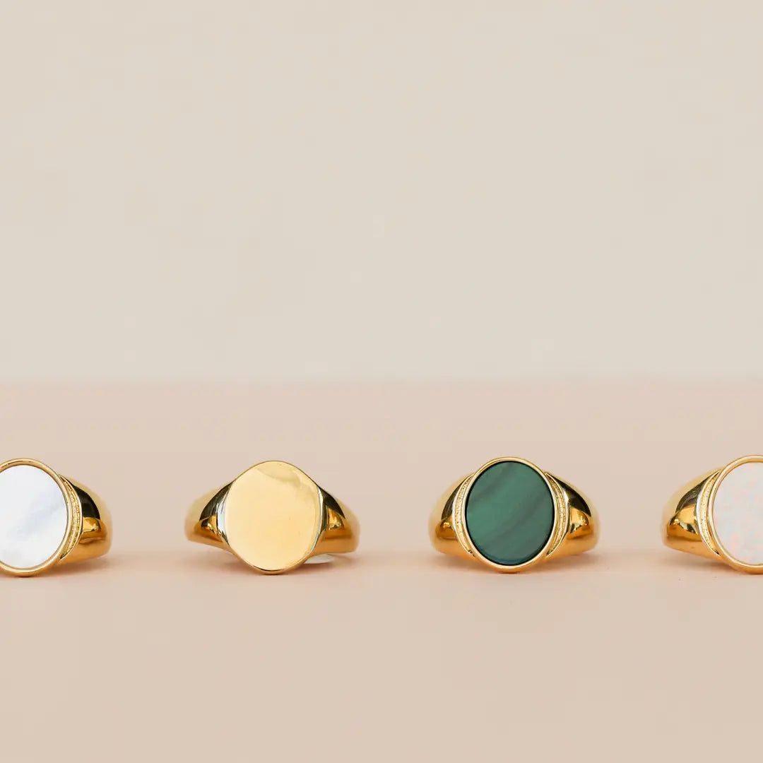 Four JaxKelly Mother of Pearl Signet Rings, each 18k gold plated with a unique color—white, gold, green, or pale pinkish-white—are displayed on a soft beige background.