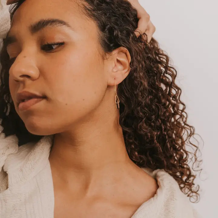 A person with curly hair wears the JaxKelly Gemstone Threader - Smoky Quartz earring and a cream-colored top, gazing to the side with one hand on their head against a plain background.