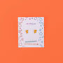 A pair of Pizza Enamel Flat Back Kids Earrings on a JaxKelly card with colorful geometric shapes, against a bright orange background. The card states they are hypoallergenic and kid-friendly—perfect for sensitive ears.