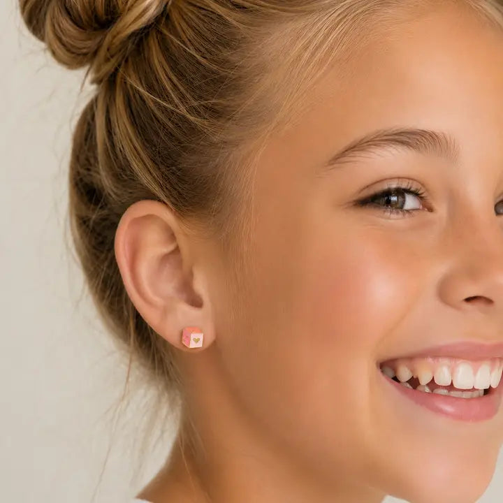 A smiling young girl with blonde hair in a bun, wearing JaxKelly Pink Dice Enamel Flat Back Kids Earrings—small, hypoallergenic earrings perfect for sensitive ears.