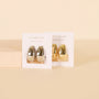Two pairs of chunky statement hoop earrings, including the Silver Bold Drop Earring, feature sterling silver posts; one pair is rhodium plated and the other gold, both displayed on white JaxKelly cards against a neutral beige background.