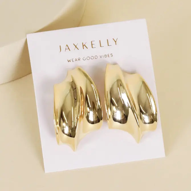 A pair of Gold Bold Wave sculptural stud earrings by JaxKelly, with 18k gold plating and a modern abstract wave design, displayed on a white card printed with JaxKelly and "Wear Good Vibes" in gold letters.