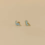 Dinosaur Enamel Flat Back Kids Earrings—two small dinosaur-shaped studs (brontosaurus and T-rex) in light blue with gold outlines, hypoallergenic for sensitive ears. Shown on a plain beige background.