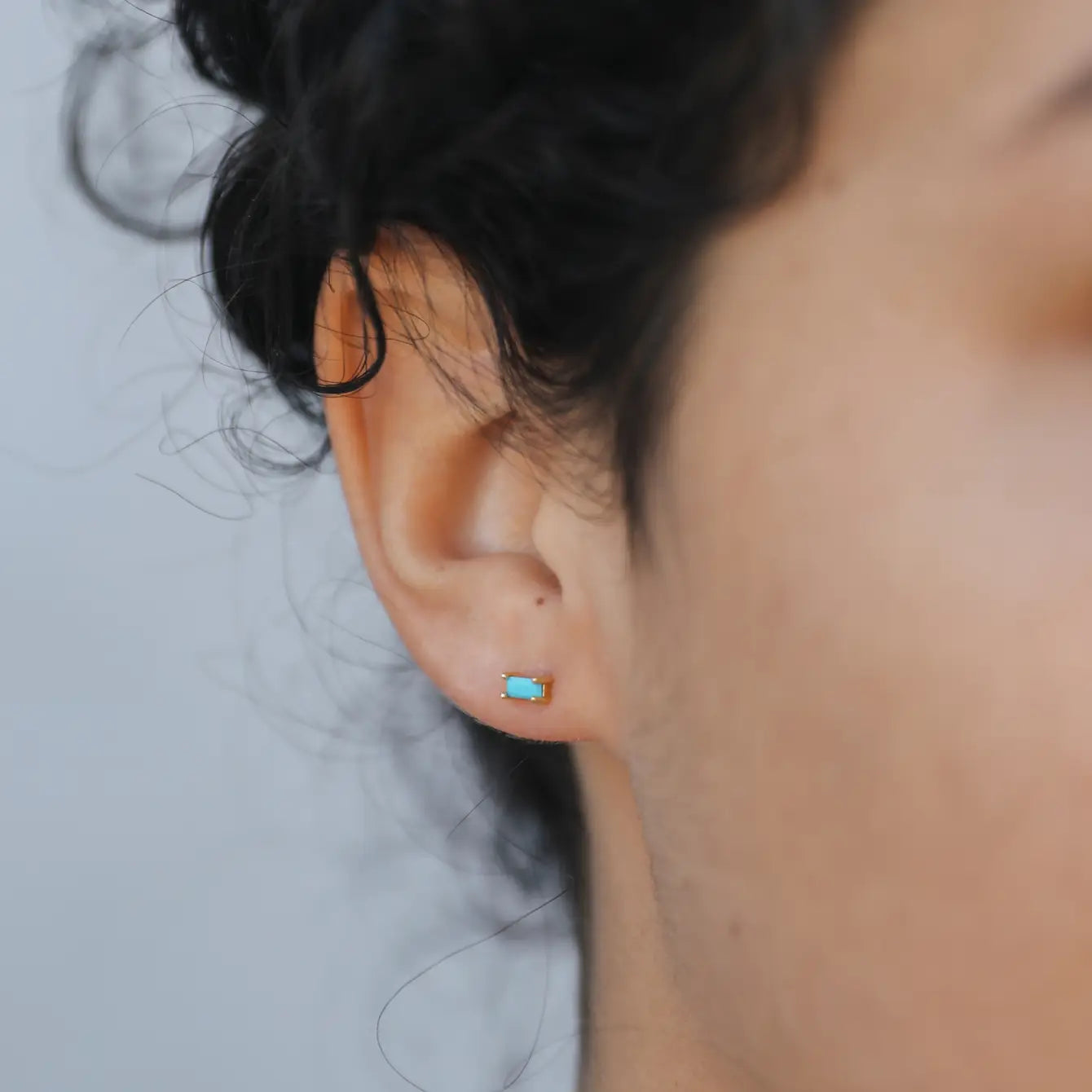Close-up of a person's ear wearing the Turquoise Baguette stud earring with sterling silver posts. The turquoise rectangular earring stands out against dark curly hair and a plain light background.