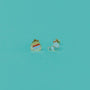Two small, colorful Bookworm Enamel Flat Back Kids Earrings on a turquoise background—one shaped like pancakes with syrup, the other a blue frog with orange eyes. Made from surgical-grade stainless steel for sensitive ears.