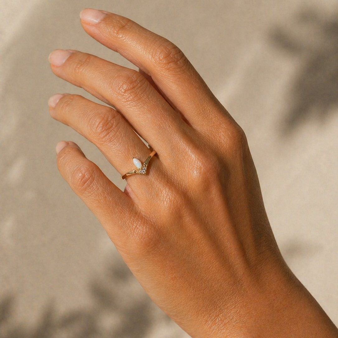 A hand with a light tan complexion wears the JaxKelly Floating Gemstone Ring - White Opal, featuring an elegant teardrop-shaped lab-grown opal and small clear stones, set against a soft beige background with subtle shadows.