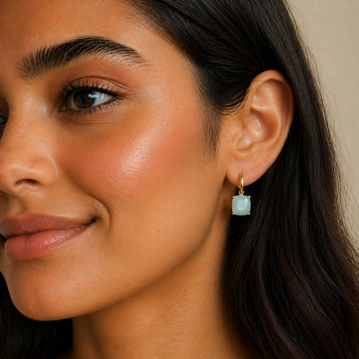 A close-up of a woman with glowing skin and long dark hair, smiling softly while wearing JaxKelly’s Gemstone Mini Drop - Amazonite earrings. The tarnish-free, 18K gold plated jewelry adds an elegant touch.