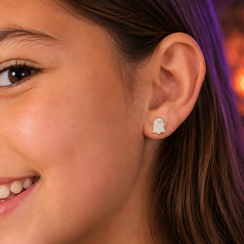 A close-up of a smiling girl’s face, focusing on her ear, which is adorned with a small, adorable JaxKelly Ghost Enamel Flat Back Kids Earring—perfect for sensitive ears. The softly blurred background features hints of purple and orange tones.