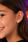 A close-up of a smiling girl’s face, focusing on her ear, which is adorned with a small, adorable JaxKelly Ghost Enamel Flat Back Kids Earring—perfect for sensitive ears. The softly blurred background features hints of purple and orange tones.