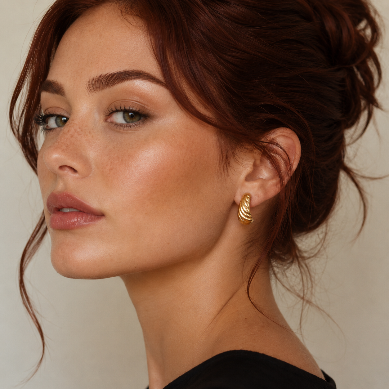 A woman with fair skin and auburn hair styled up looks over her shoulder, wearing JaxKelly’s Gold Bold Swirl Earrings. She has defined brows, soft pink lips, subtle makeup, and a black top set against a neutral background.
