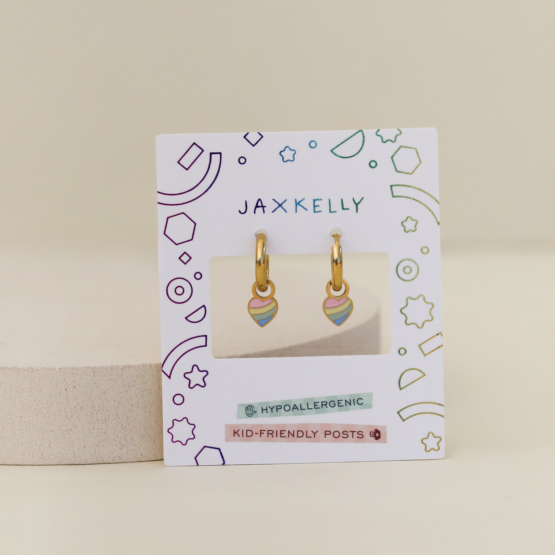 The Heart Enamel Charm Hoop earrings are gold hoops with rainbow heart charms for sensitive ears, shown on a white JaxKelly card with colorful geometric and celestial designs. The hypoallergenic pair is also kid-friendly.