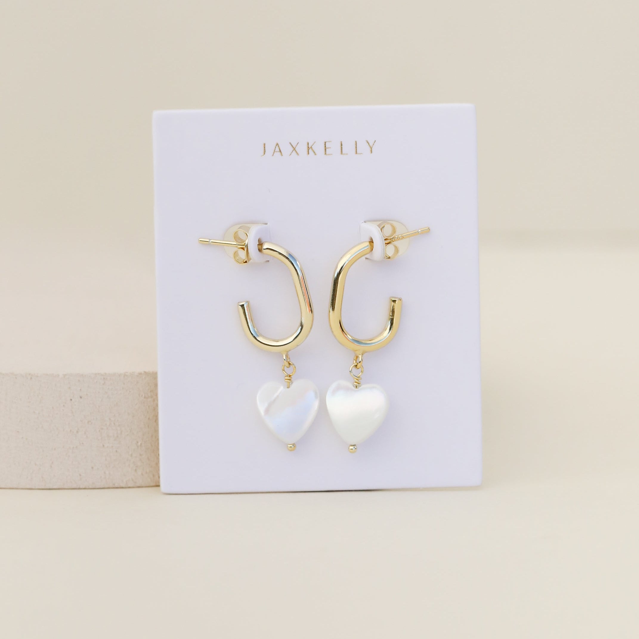 The Heart Drop - Mother of Pearl are tarnish-free gold hoop earrings with dangling white heart-shaped charms, shown on a white JaxKelly card against a beige background.