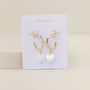 The Heart Drop - Mother of Pearl are tarnish-free gold hoop earrings with dangling white heart-shaped charms, shown on a white JaxKelly card against a beige background.