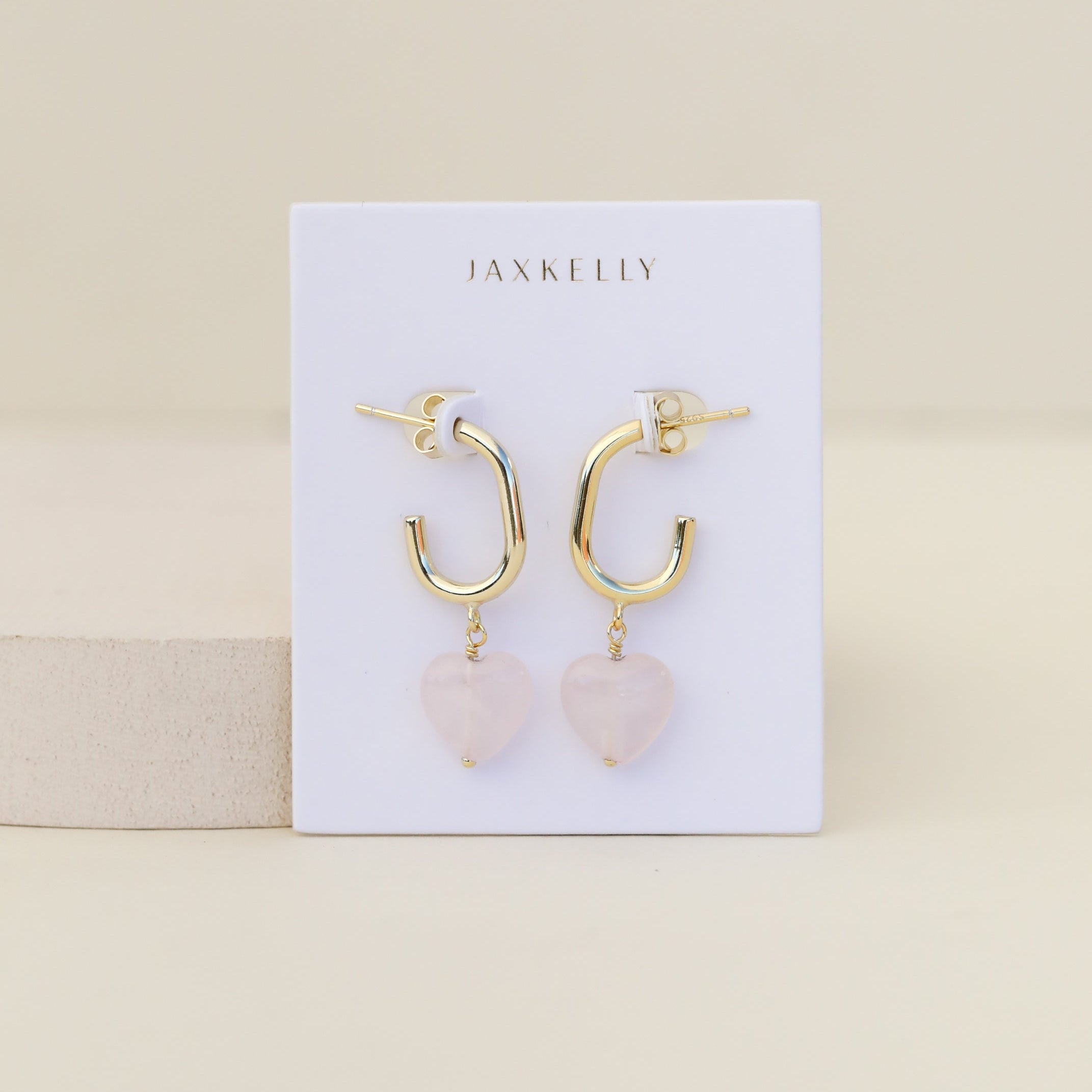 The Heart Drop - Rose Quartz earrings feature gold hoops with rose quartz heart pendants, displayed on a white JaxKelly card against a beige background. These chic accessories are tarnish free for lasting shine.