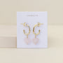 The Heart Drop - Rose Quartz earrings feature gold hoops with rose quartz heart pendants, displayed on a white JaxKelly card against a beige background. These chic accessories are tarnish free for lasting shine.