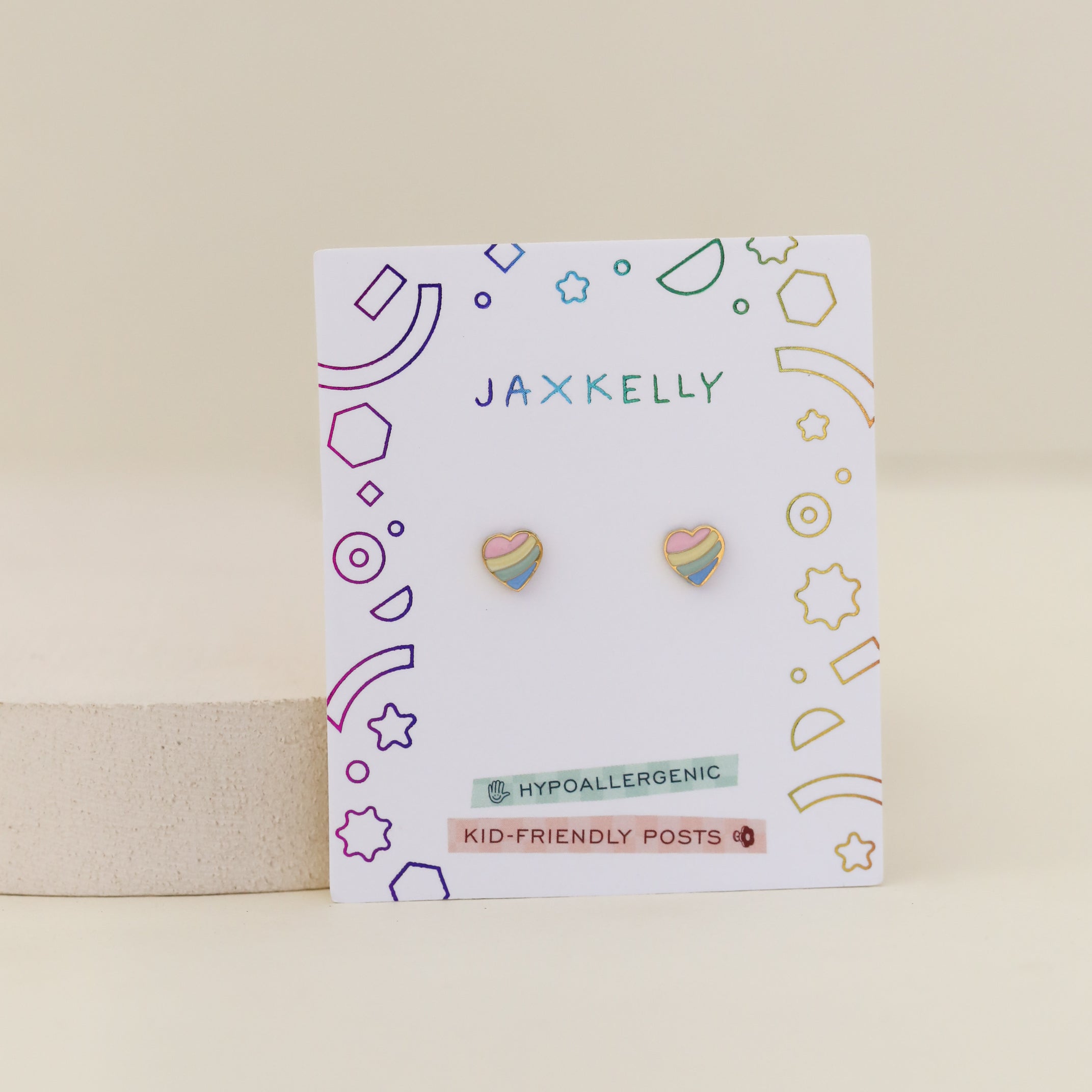 The Heart Enamel Flat Back Kids Earrings are small, heart-shaped studs in pastel colors with hypoallergenic, kid-friendly posts—perfect for sensitive ears and featuring colorful geometric designs on a white JaxKelly card.