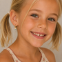 A young blonde girl in pigtails smiles at the camera, wearing JaxKelly's Heart Enamel Charm Hoop earrings—hypoallergenic and perfect for kids’ sensitive ears. She pairs them with a cream floral embroidered top against a neutral backdrop.
