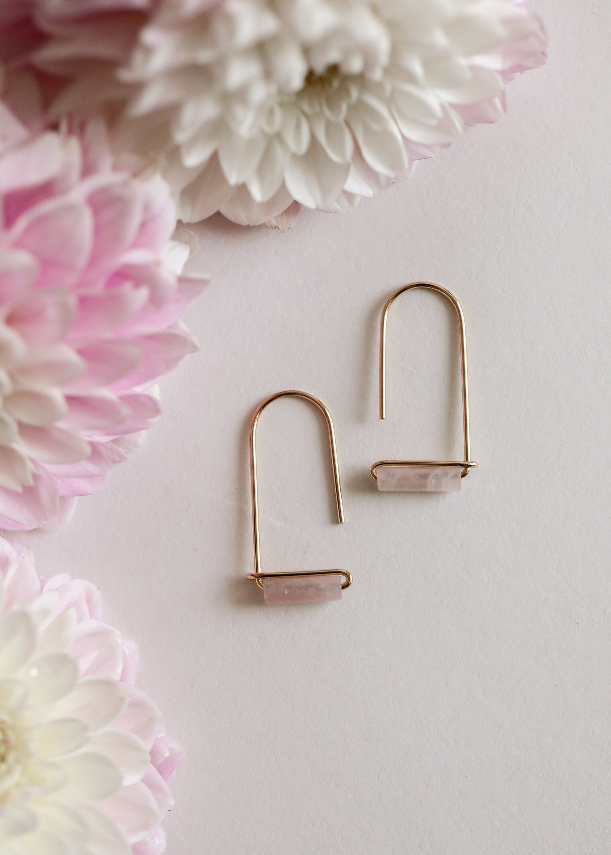 Two Rose Quartz Gemstone Drop - Love earrings by JaxKelly with minimalist gold plating and rectangular pink stones are displayed on a light surface among soft pink and white flowers, adding an elegant touch of gemstone jewelry.