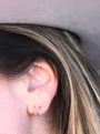 Close-up of a person's ear wearing two JaxKelly Minimalist Spiral gold plated hoops. The person has light skin and straight brown hair with blonde highlights; the background is blurred and gray.