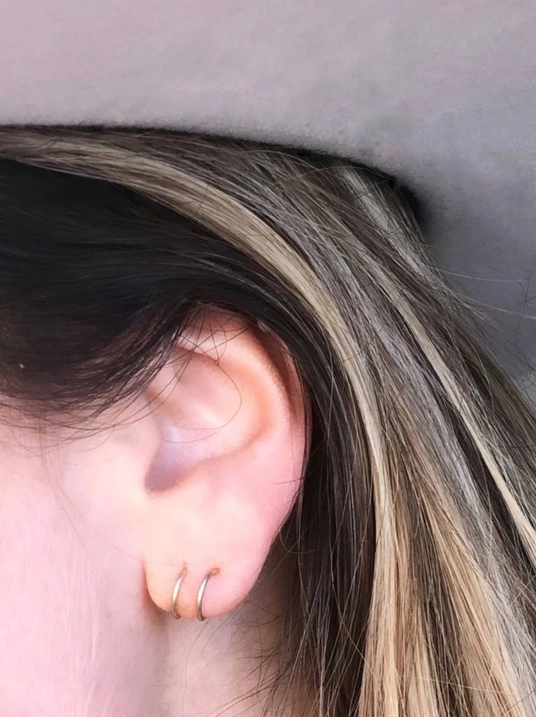 Close-up of a person's ear wearing two JaxKelly Minimalist Spiral gold plated hoops. The person has light skin and straight brown hair with blonde highlights; the background is blurred and gray.