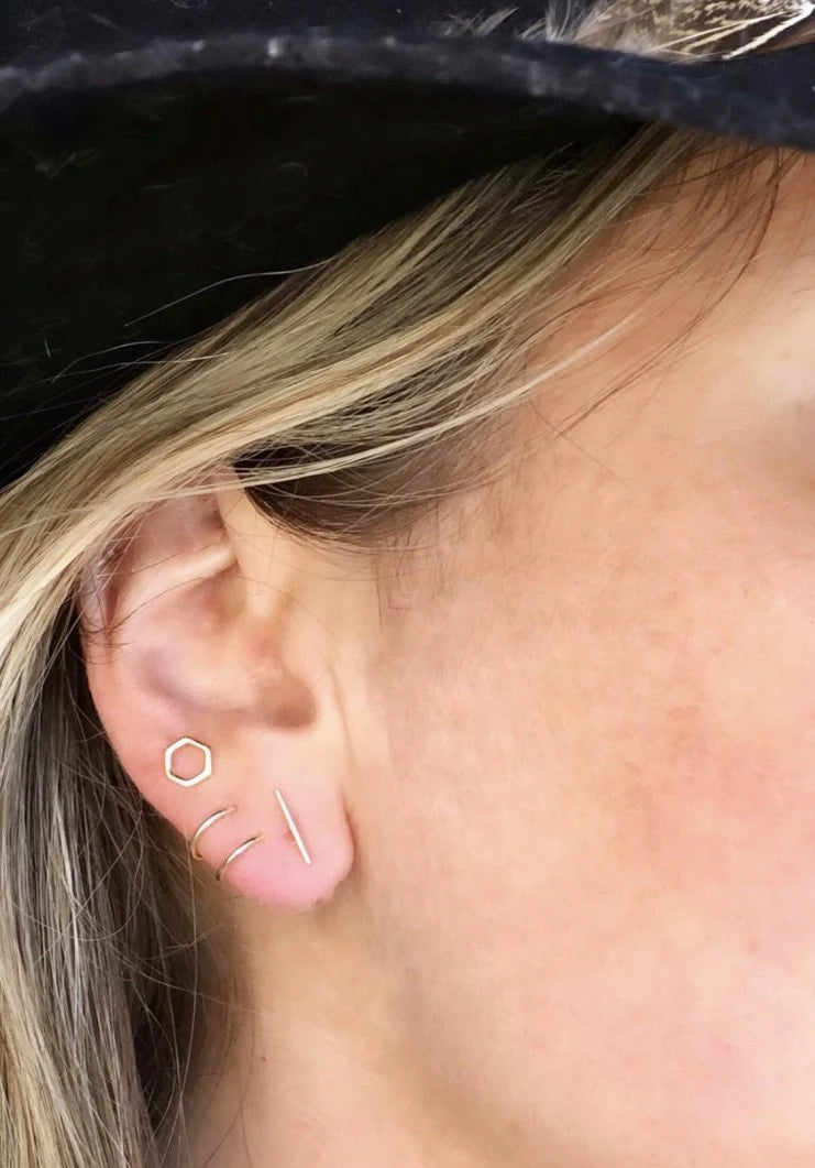 A close-up of a person's ear wearing the JaxKelly Minimalist Spiral gold plated earrings: a hexagon stud, vertical bar, curved layered hoop, and horizontal bar. The person has blonde hair and is wearing a black hat.