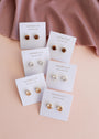 A pair of Round CZ - Coffee - Gold Earrings by JaxKelly is displayed on a white brand card, arranged on a pink surface with a mauve cloth in the top corner.