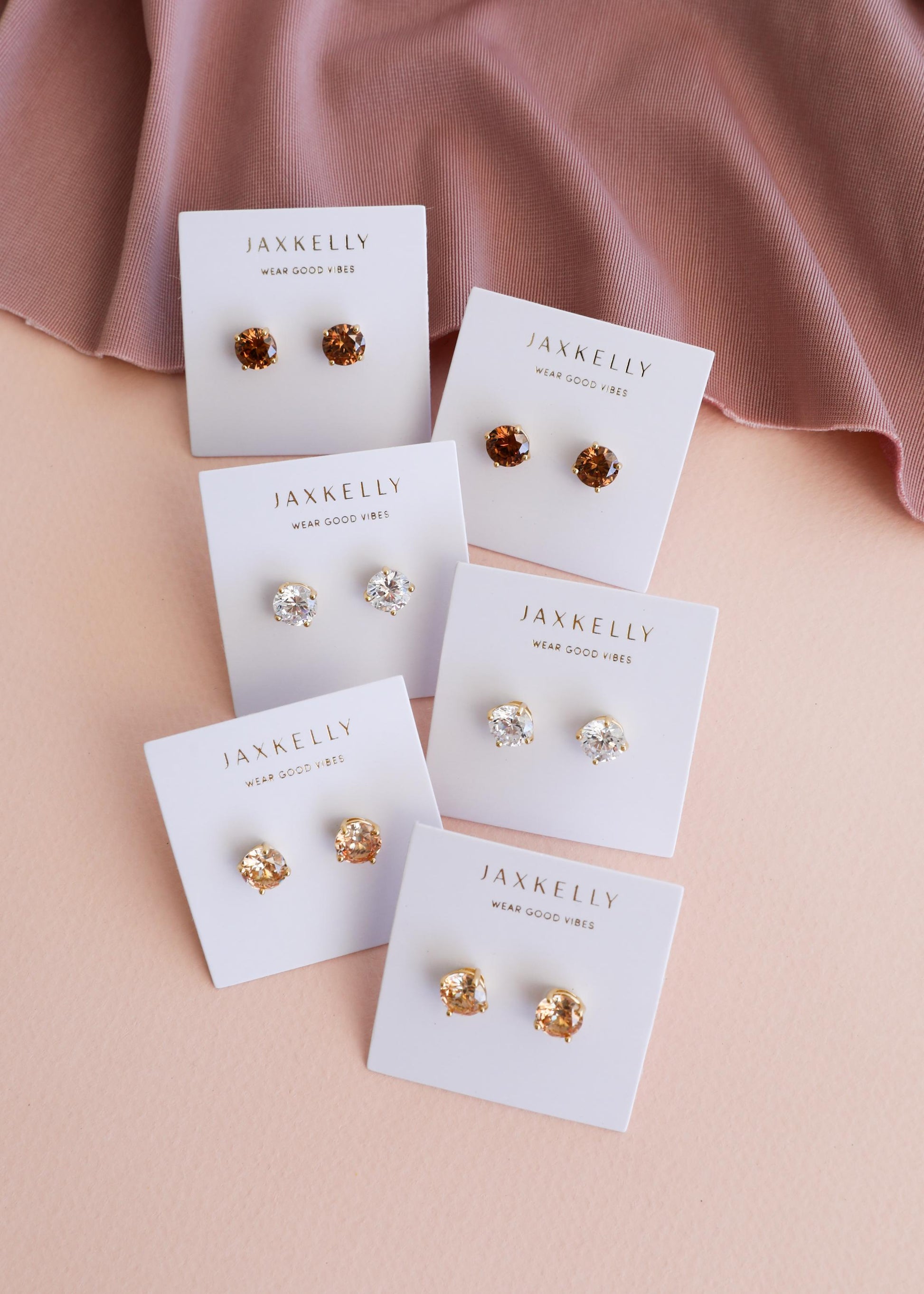 A pair of Round CZ - Coffee - Gold Earrings by JaxKelly is displayed on a white brand card, arranged on a pink surface with a mauve cloth in the top corner.