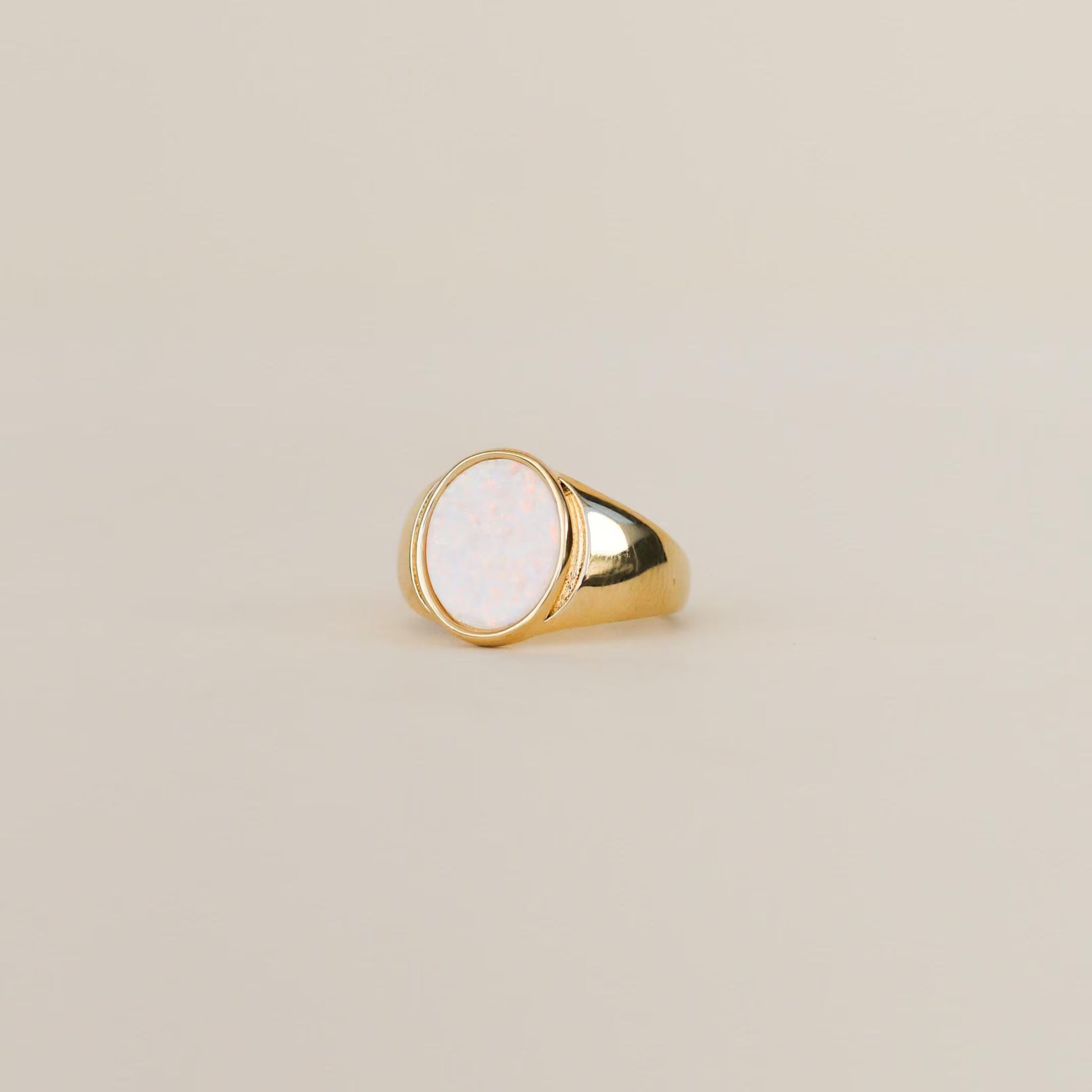 The White Opal Signet Ring by JaxKelly features a large oval white opal set in 18k gold plating, showcased on a simple beige background.