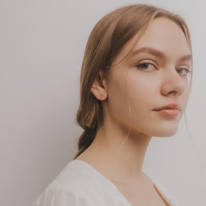 A young woman with light brown hair tied back, wearing a white top and JaxKelly's Cross Stud 18k gold plated earrings, gazes calmly at the camera against a plain light background.