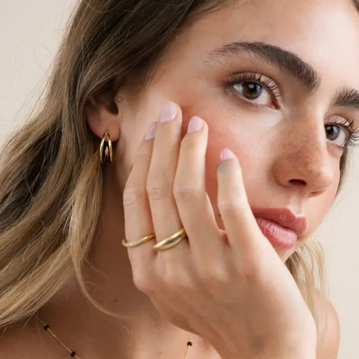 A young woman with natural makeup and light brown hair touches her face, wearing JaxKelly's Illusion Double Hoop - Gold Earrings, a gold ring, a delicate necklace, and showing off her freckles.