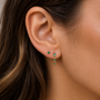 A close-up of a woman’s ear showcases the JaxKelly Mini Baguette Drop Earrings - Emerald CZ: a gold-plated stud and a dangling hypoallergenic earring with rectangular and square green stones, framed by her wavy brown hair.