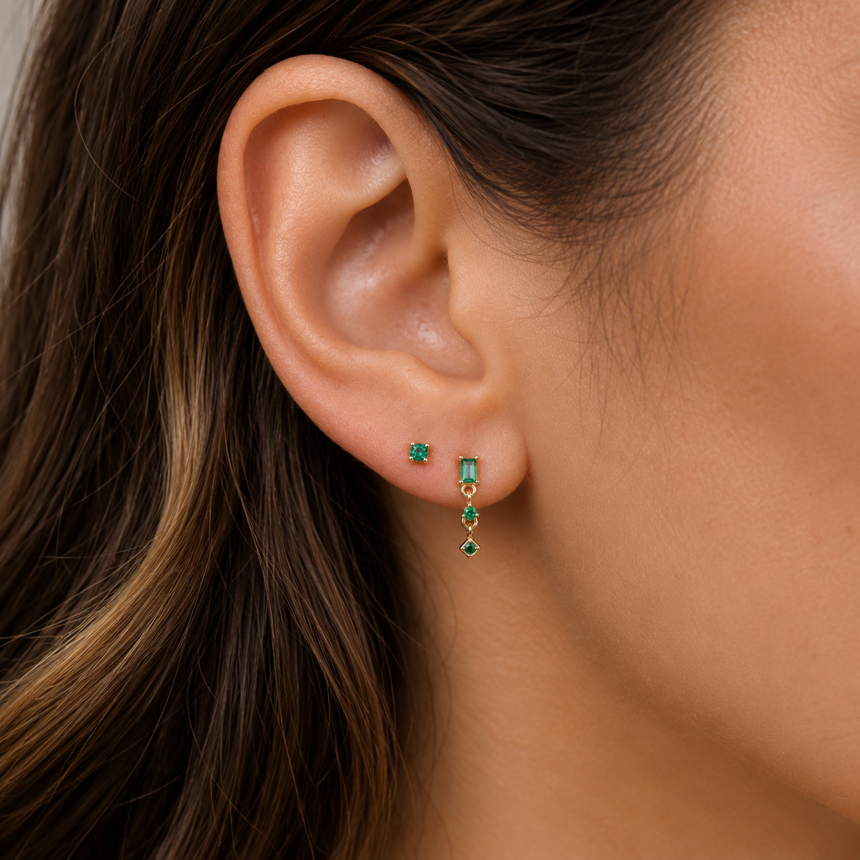 A close-up of a woman’s ear showcases the JaxKelly Mini Baguette Drop Earrings - Emerald CZ: a gold-plated stud and a dangling hypoallergenic earring with rectangular and square green stones, framed by her wavy brown hair.
