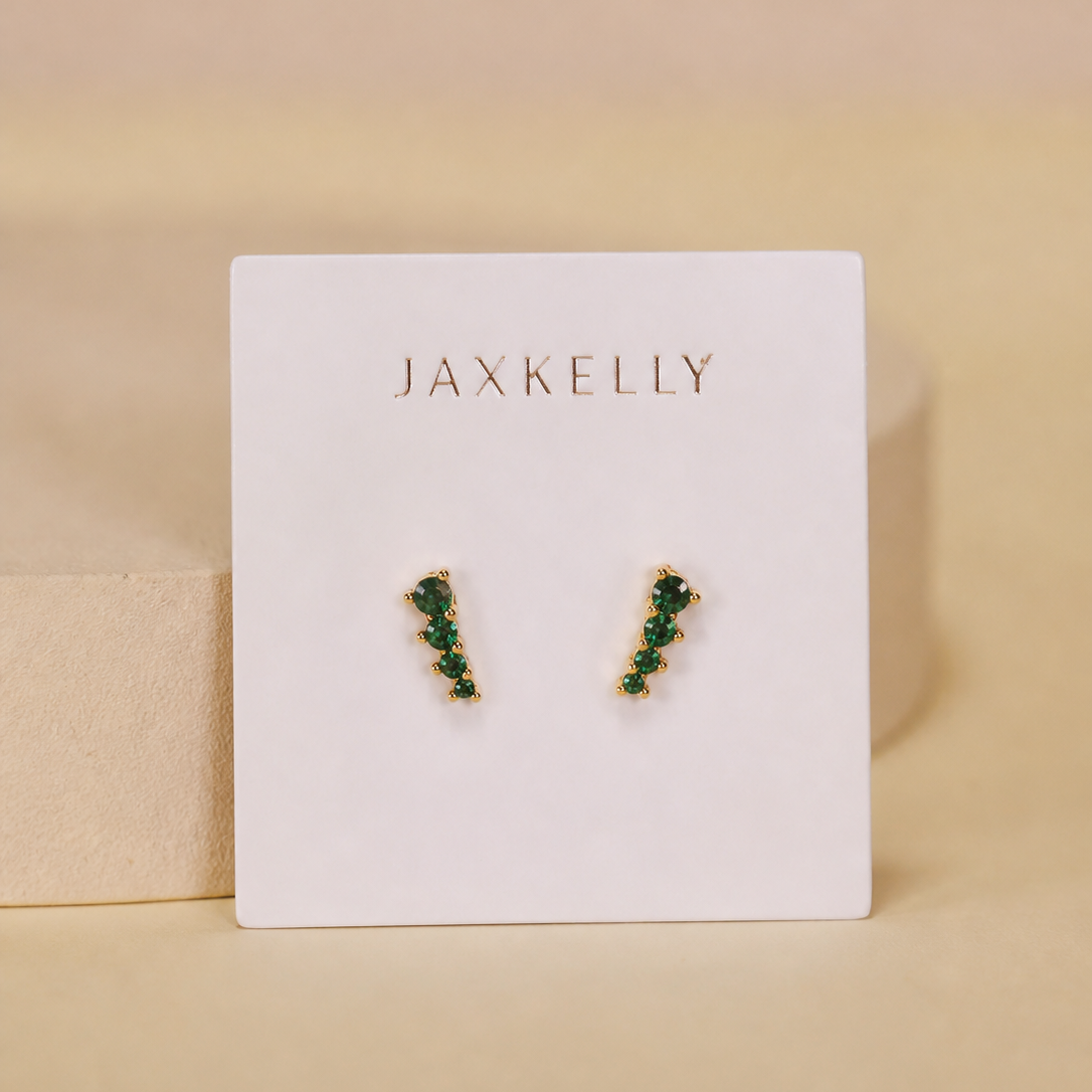 A pair of JaxKelly Mini Crawler - Emerald CZ - Gold Earrings features 18K gold plating and small green stones in a vertical line on a white branded card with gold text, set against a tan background. Hypoallergenic for sensitive ears.