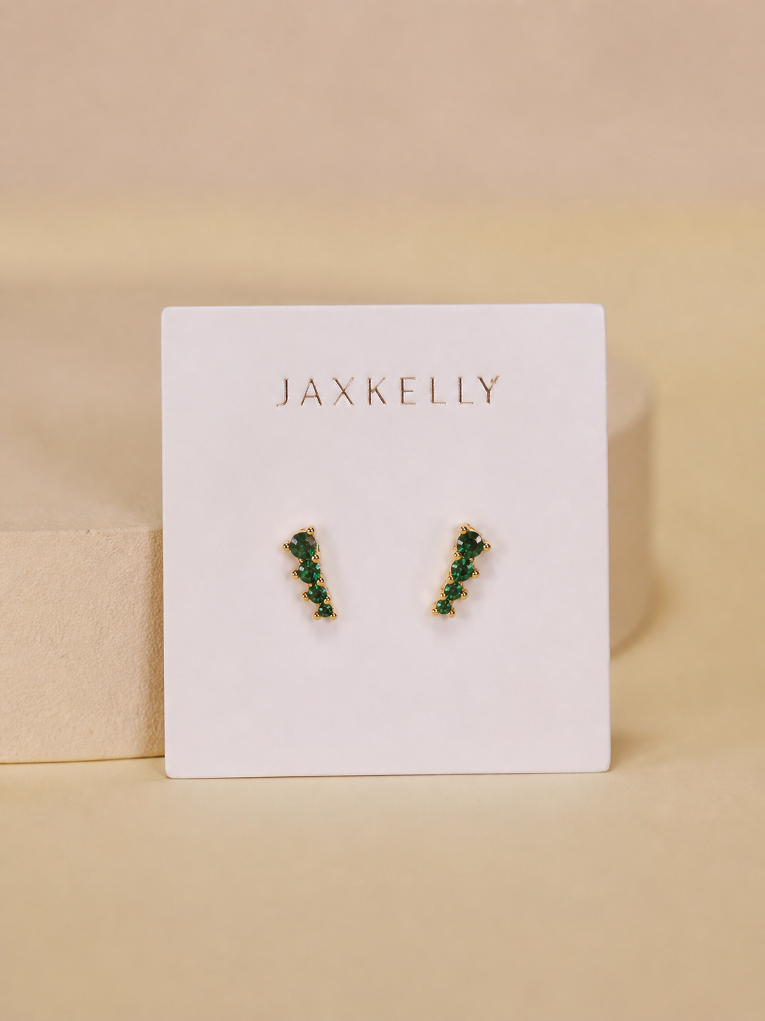 A pair of JaxKelly Mini Crawler - Emerald CZ - Gold Earrings features 18K gold plating and small green stones in a vertical line on a white branded card with gold text, set against a tan background. Hypoallergenic for sensitive ears.